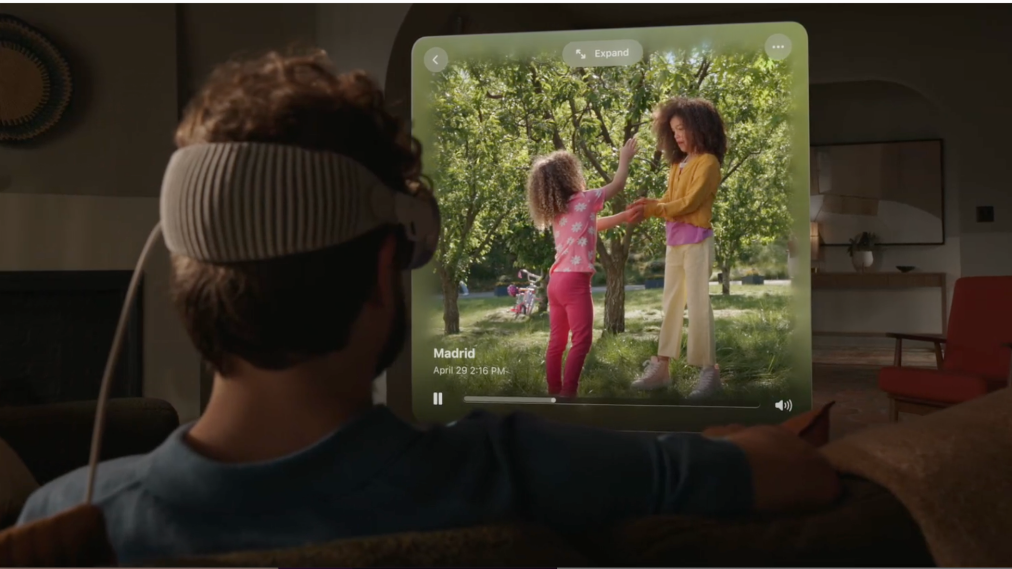 A white curly-haired man wearing the Apple Vision Pro is seen from the back. He is watching video of two girls (presumably his daughters) on a screen projected in front of him